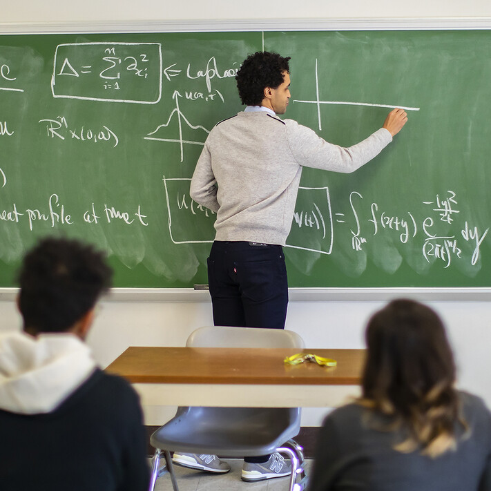 graduate students in a classroom with one writing on a chalkboard